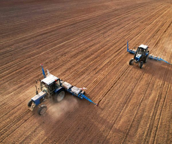 Tractor sowing seed on plowed field. Sowing seeds of corn and sunflower. Blue Tractor with disk harrow on plowing field. Seeding machinery on farm field. Seed sowing in farmland, aerial view..